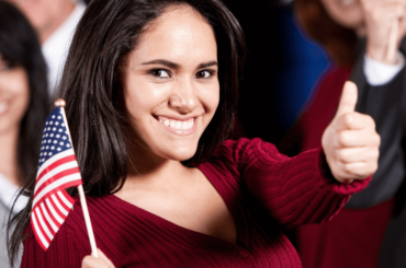 woman in red t-shirt waving a US flag in one hand and thumbs up with the other hand